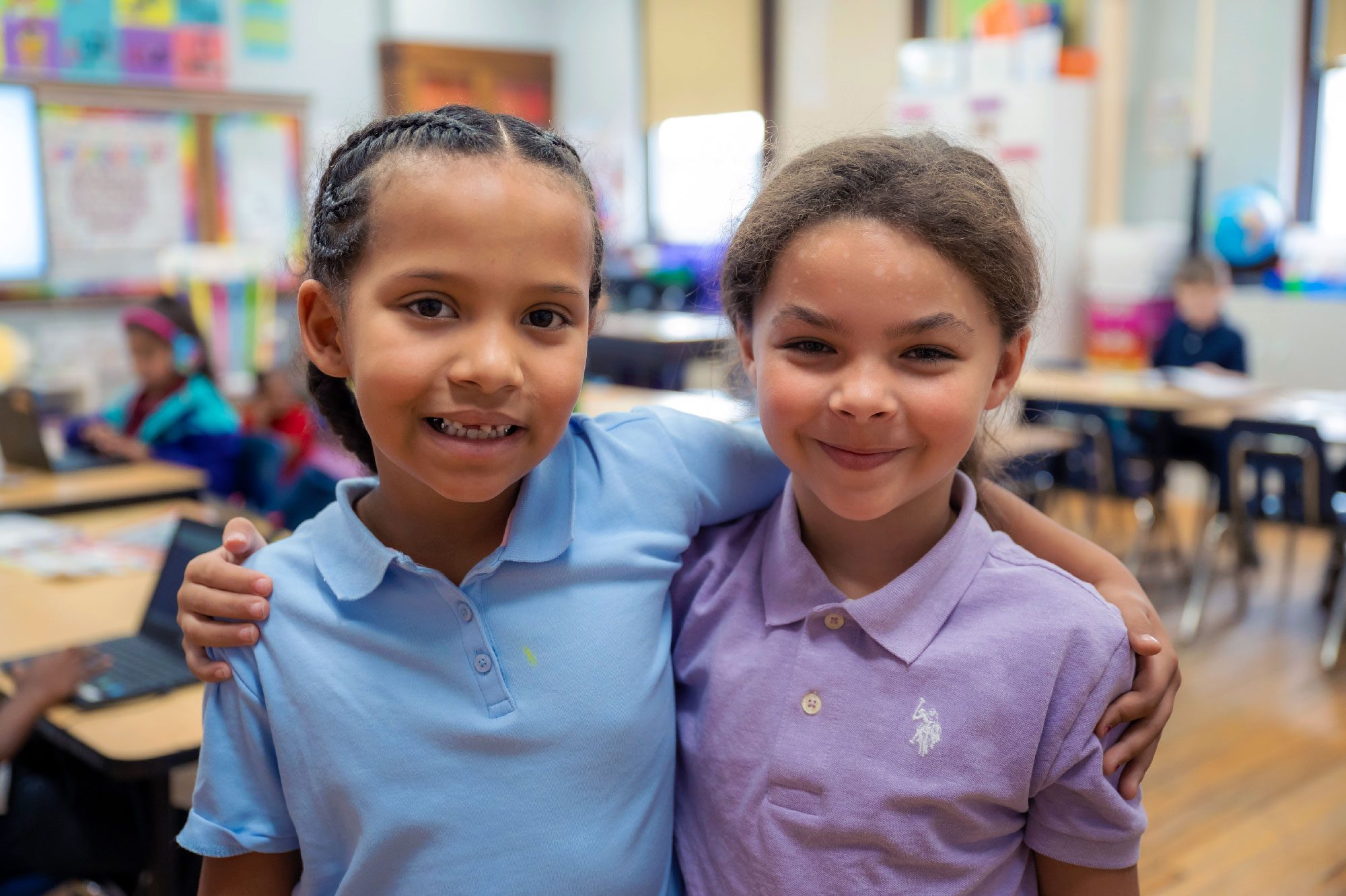 Two smiling students in school uniform polos standing arm in arm in a classroom.